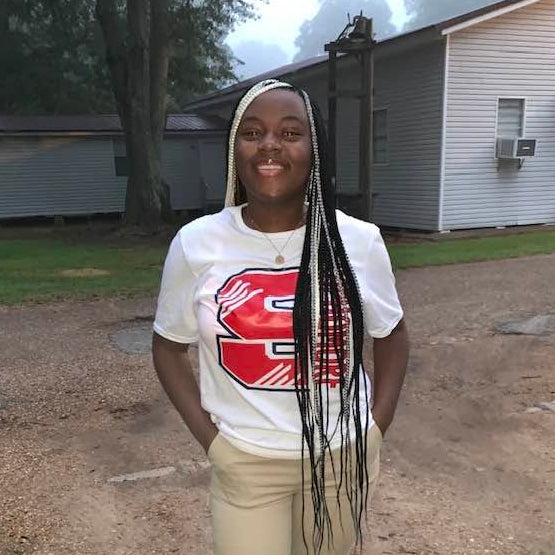 Person wearing a white t-shirt with a red logo and beige pants standing on a dirt road.